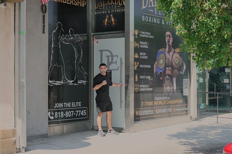 Artem Dalakian in a black T-shirt and shorts holding open the frosted glass front door of the gym, between window posters that read JOIN THE ELITE and TRAIN WITH A 7-TIME WORLD CHAMPION