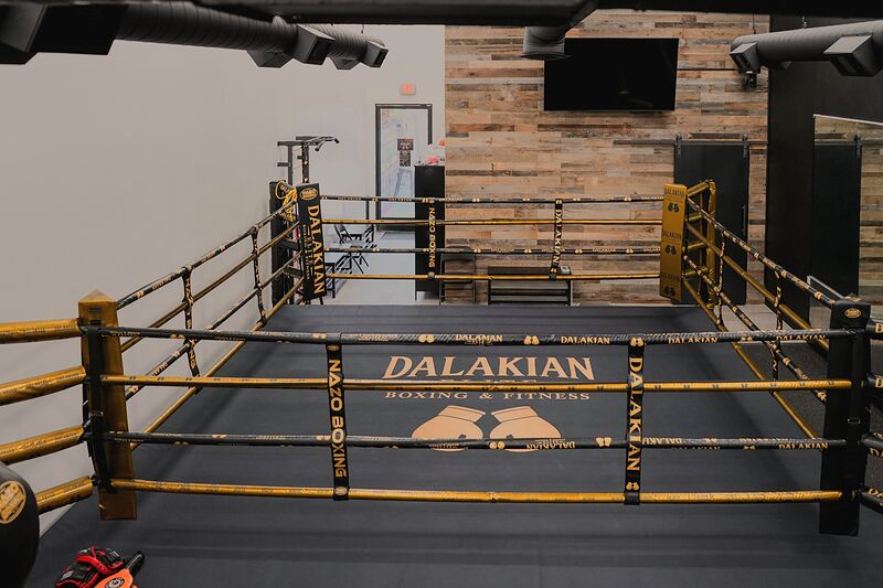 The boxing ring seen from a corner: black canvas printed DALAKIAN BOXING & FITNESS with two gold gloves, black and gold ropes and gold corner posts