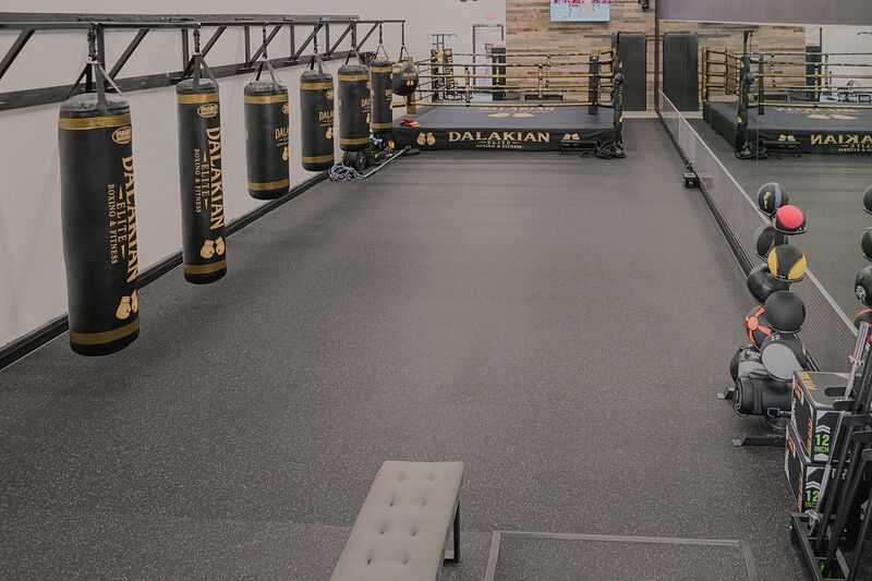 A row of black heavy bags with gold trim hanging from a steel rack along a white wall, medicine balls on a rack at the right and the ring at the far end