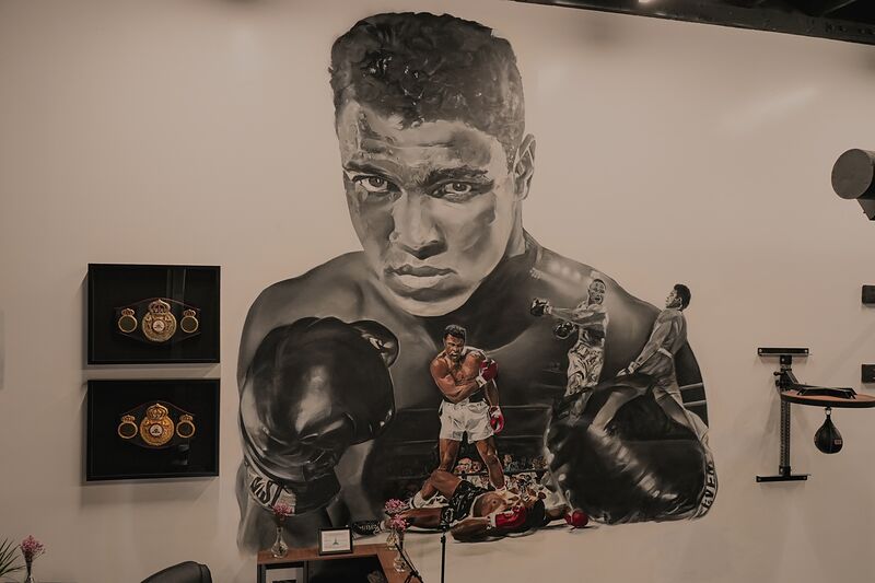 Two WBA championship belts in black frames on a white wall, beside a large black and white mural of Muhammad Ali