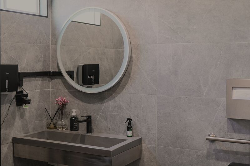 Restroom with grey tile, a round lit mirror, a stainless steel sink and a grab bar